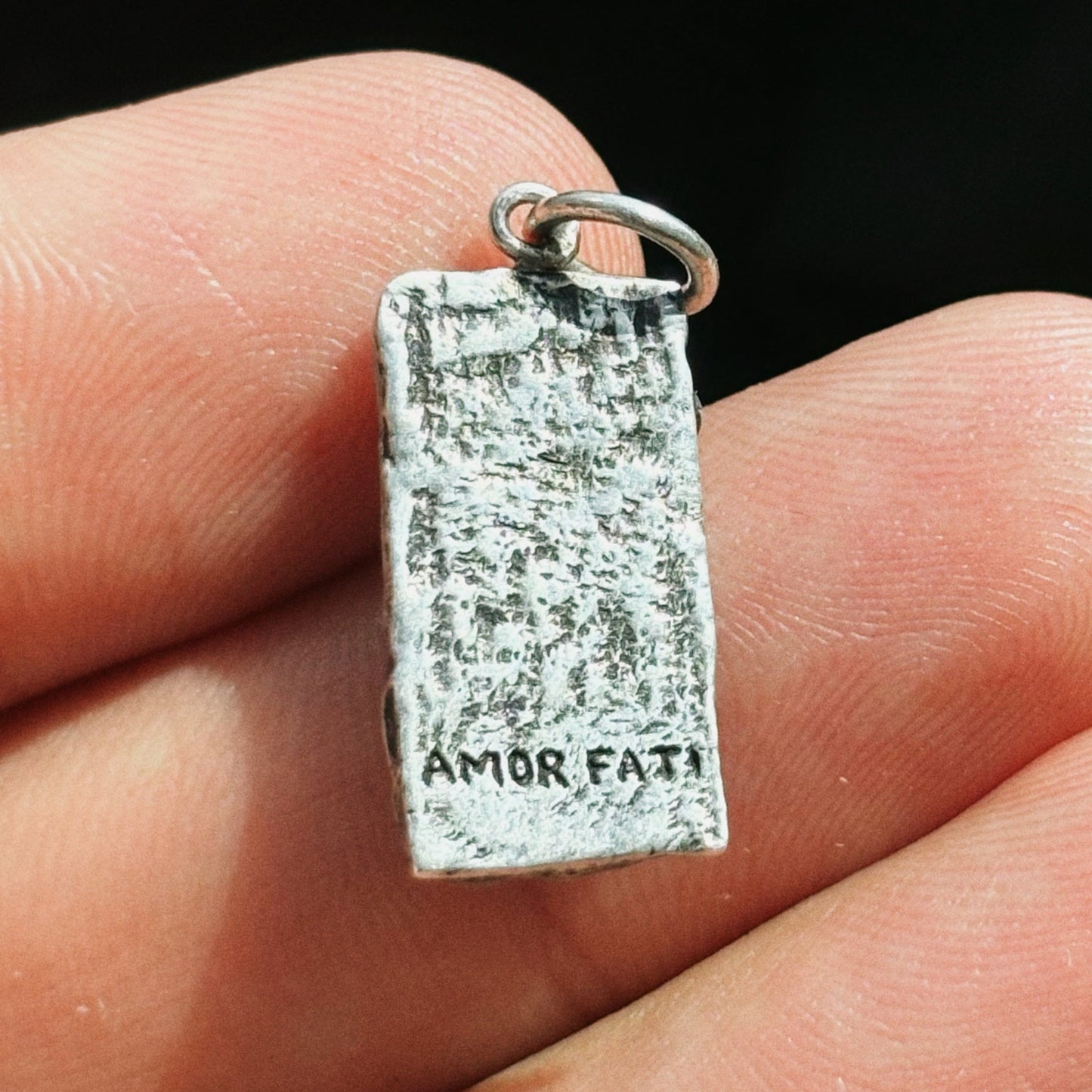 Silver 'Amor Fati' pendant held in a hand against a dark background