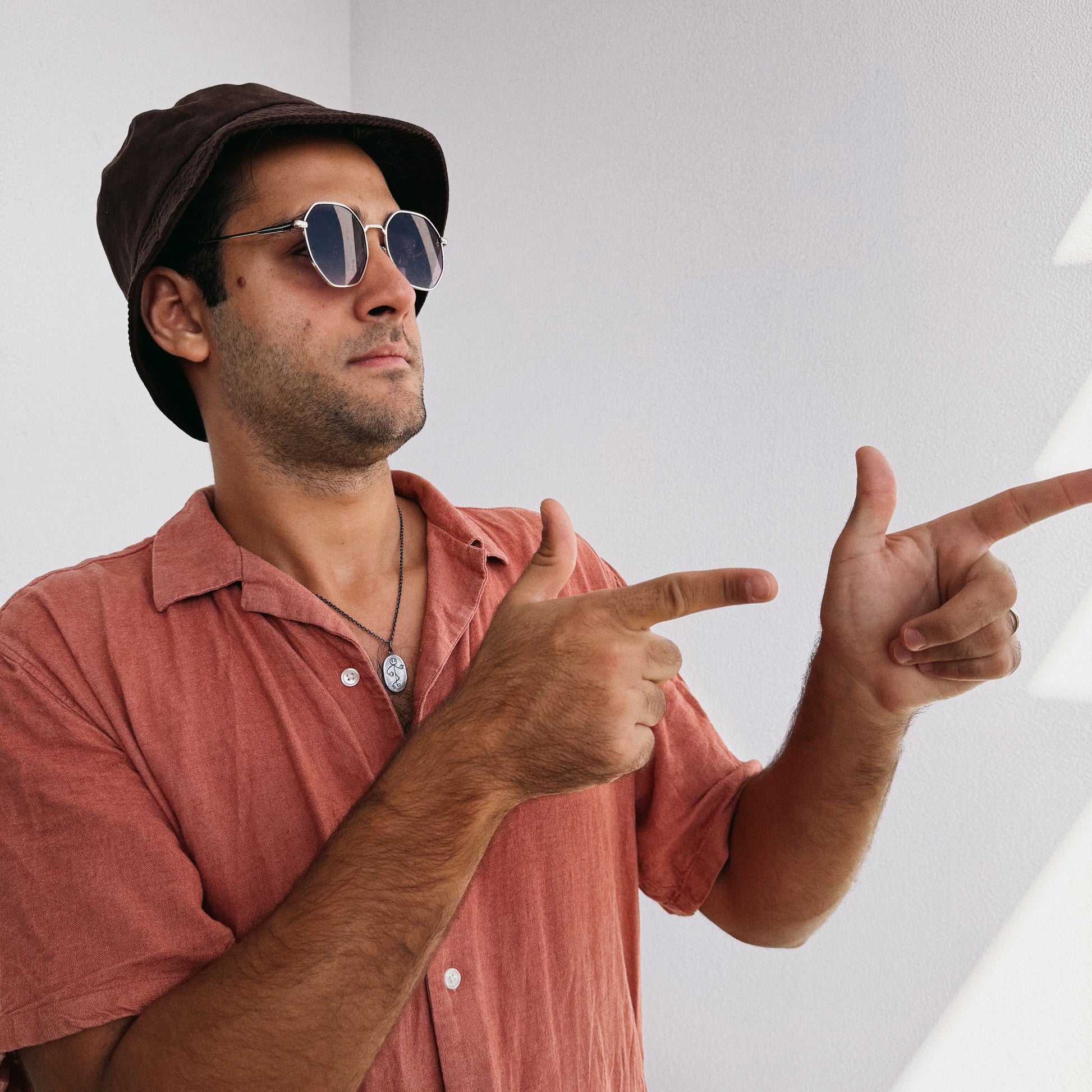 Man wearing a brown cap and sunglasses, pointing towards the camera against a white background