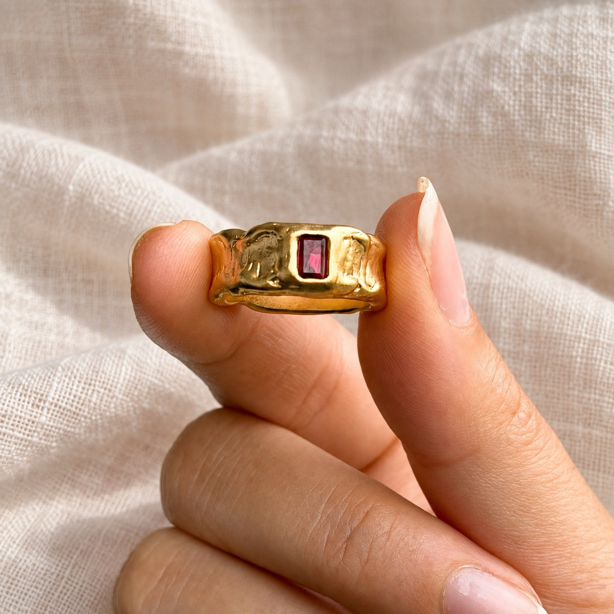 A close-up of a hand holding a textured gold ring with a rectangular red gemstone, set against a soft, draped beige linen fabric background.