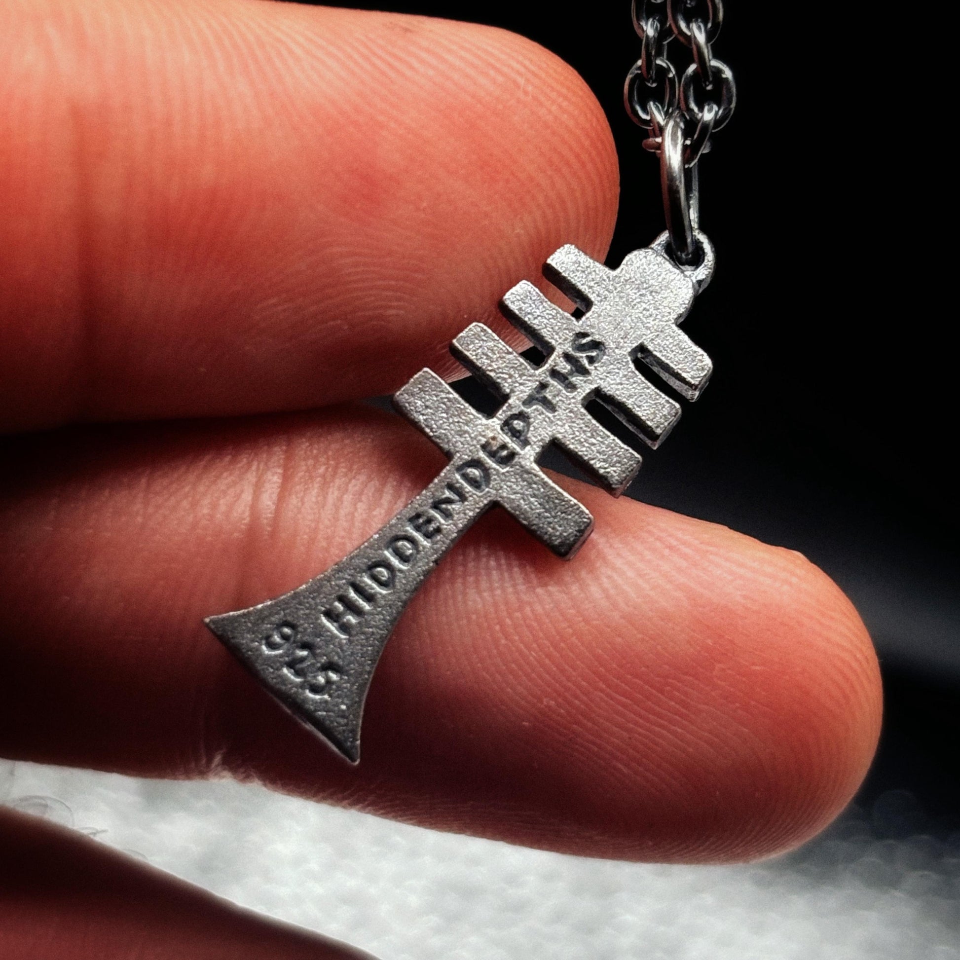 Silver cross-shaped pendant with engraved text held between fingers against a dark background