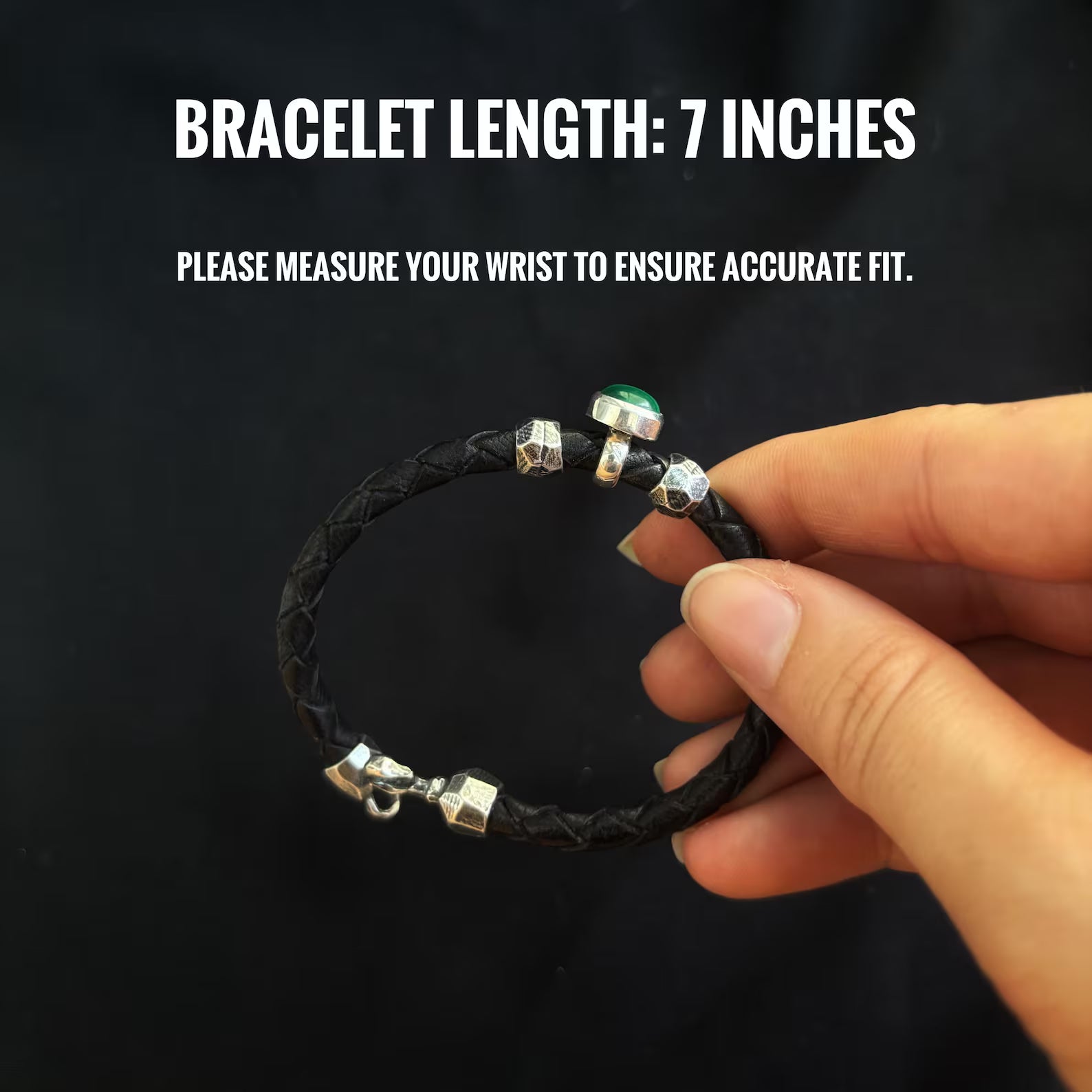 Black braided bracelet with silver and green accents held by a hand against a black background.