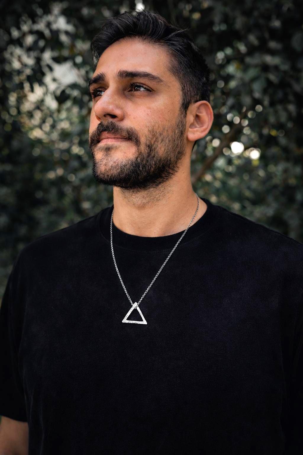 Man wearing a black t-shirt with a triangle necklace against a blurred green background